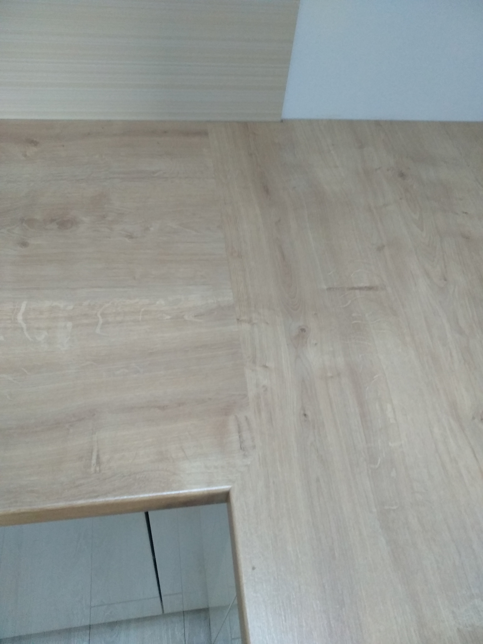 Fragment drewnianej podłogi z widocznym łączeniem paneli i fragmentem ościeżnicy drzwiowej w dolnym lewym rogu, ukazujący wnętrze z białymi płytkami.