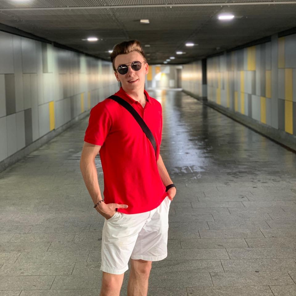 Młody mężczyzna w czerwonej koszulce polo i białych szortach pozuje w długim, oświetlonym tunelu z szarymi ścianami i żółtymi akcentami. Ma na sobie okulary przeciwsłoneczne i czarną torbę na ramię.