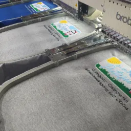 Szara koszulka umieszczona w ramie hafciarki Brother, z wyhaftowanym kolorowym logo firmy sprzątającej 'Grzegorz Ostrowski Gebäudereinigung seit 2004', widoczne igły i nici maszyny.