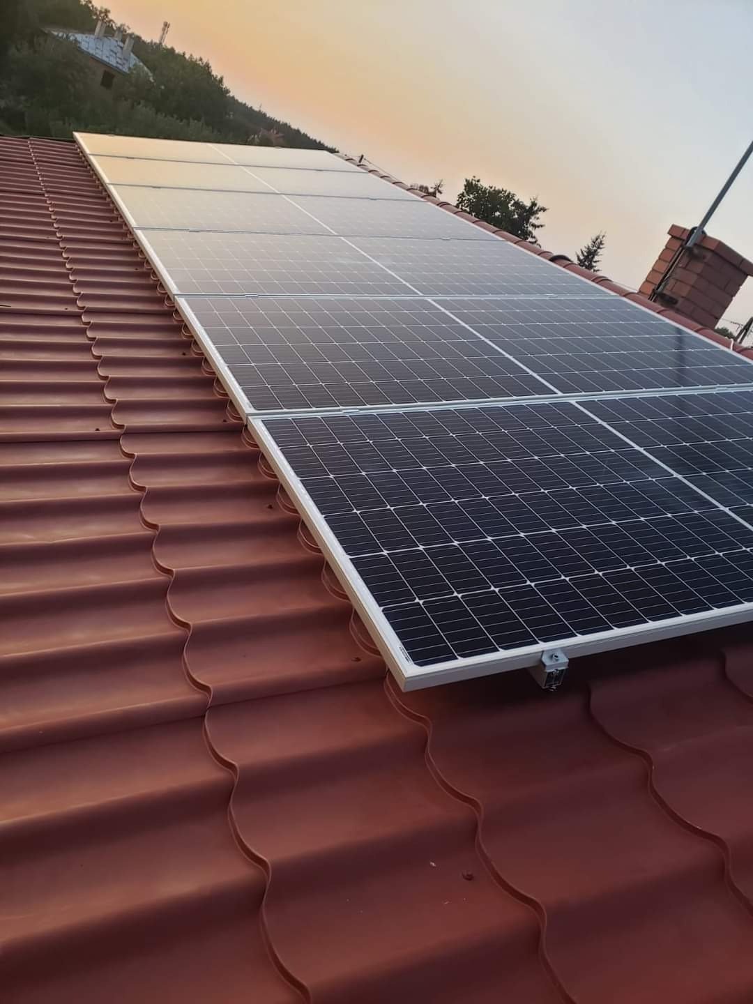Panel słoneczny z widocznym mocowaniem na czerwonej dachówce w promieniach zachodzącego słońca.
