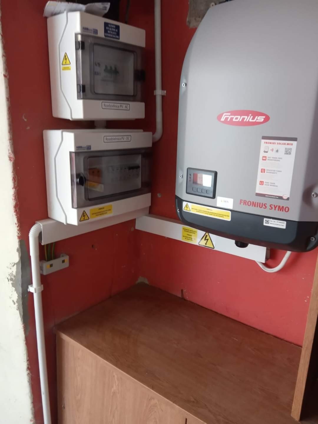 Instalacja elektryczna wewnątrz budynku z dwoma rozdzielnicami PV (AC i DC) oraz inwerterem marki Fronius SYMO, widoczne okablowanie i ostrzeżenia o napięciu.