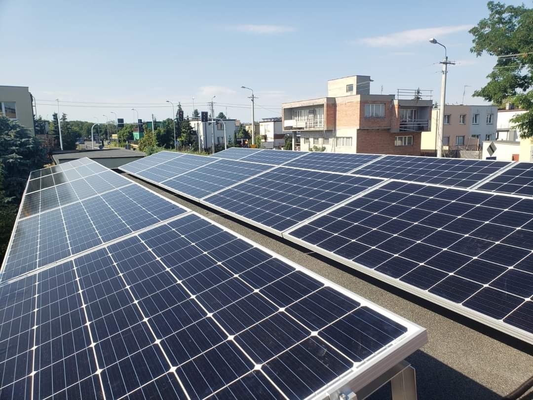 Instalacja paneli słonecznych na płaskim dachu budynku mieszkalnego, widok z góry na panele z widocznymi elementami montażowymi, w tle zabudowa miejska i błękitne niebo.