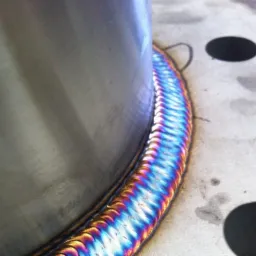 Metalowy cylinder z precyzyjnym, wielobarwnym spoiwem na krawędzi łączenia z płaską powierzchnią, widoczne otwory montażowe.