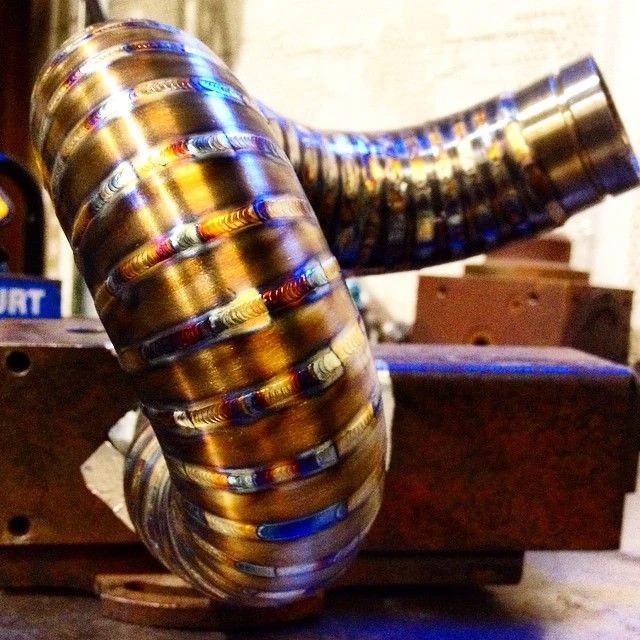 Precyzyjnie wykonane spawy na zakrzywionej rurze metalowej, widoczne tęczowe przebarwienia ciepła, zamocowanej w uchwycie tokarskim.