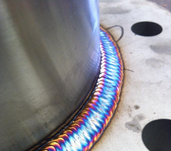 Metalowy cylinder z precyzyjnym, wielobarwnym spoiwem na krawędzi łączenia z płaską powierzchnią, widoczne otwory montażowe.