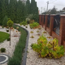 Ogród z trawnikiem, rabatami wysypanymi białym grysem, obrzeżami z ciemnej kostki, roślinami ozdobnymi i ceglanym ogrodzeniem z metalowymi przęsłami.