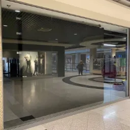 Duże, czyste okno wystawowe sklepu odzieżowego w centrum handlowym, odbijające wnętrze pasażu i sylwetkę przechodzącej kobiety.