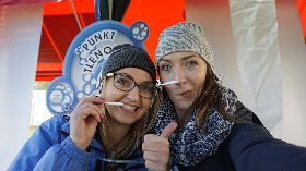 Dwie uśmiechnięte kobiety w czapkach, robiące sobie selfie z rekwizytami w kształcie wąsów na tle stoiska z napisem 'Punkt Tleno'.