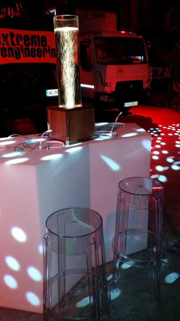 Podświetlany cylinder z dekoracją, transparentne krzesła barowe, biały stół i ciężarówka w tle podczas eventu firmowego z czerwonym oświetleniem.