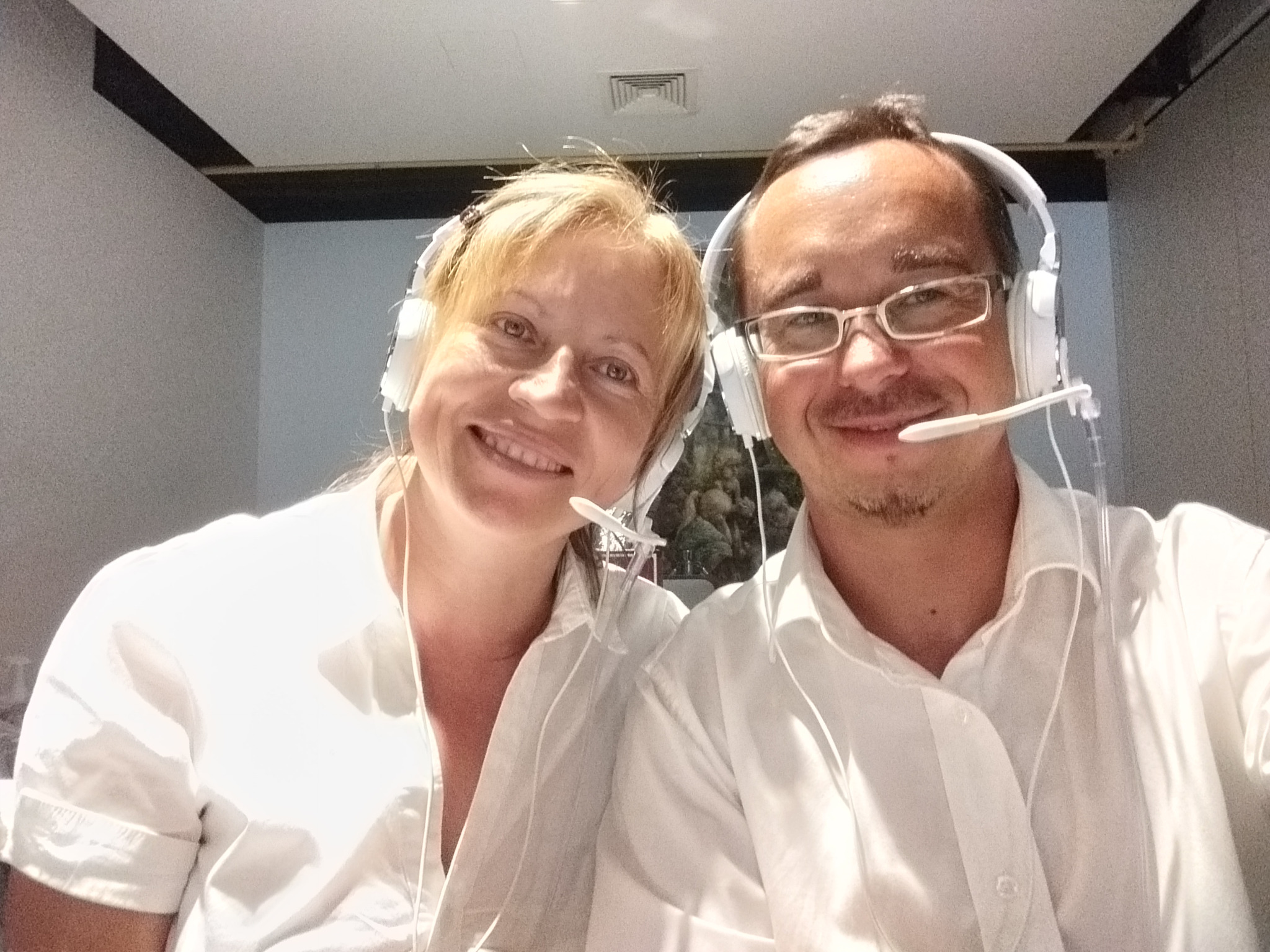 Uśmiechnięta kobieta i mężczyzna w białych koszulach i słuchawkach z mikrofonami, prawdopodobnie obsługa techniczna wydarzenia lub konsultanci telefoniczni.