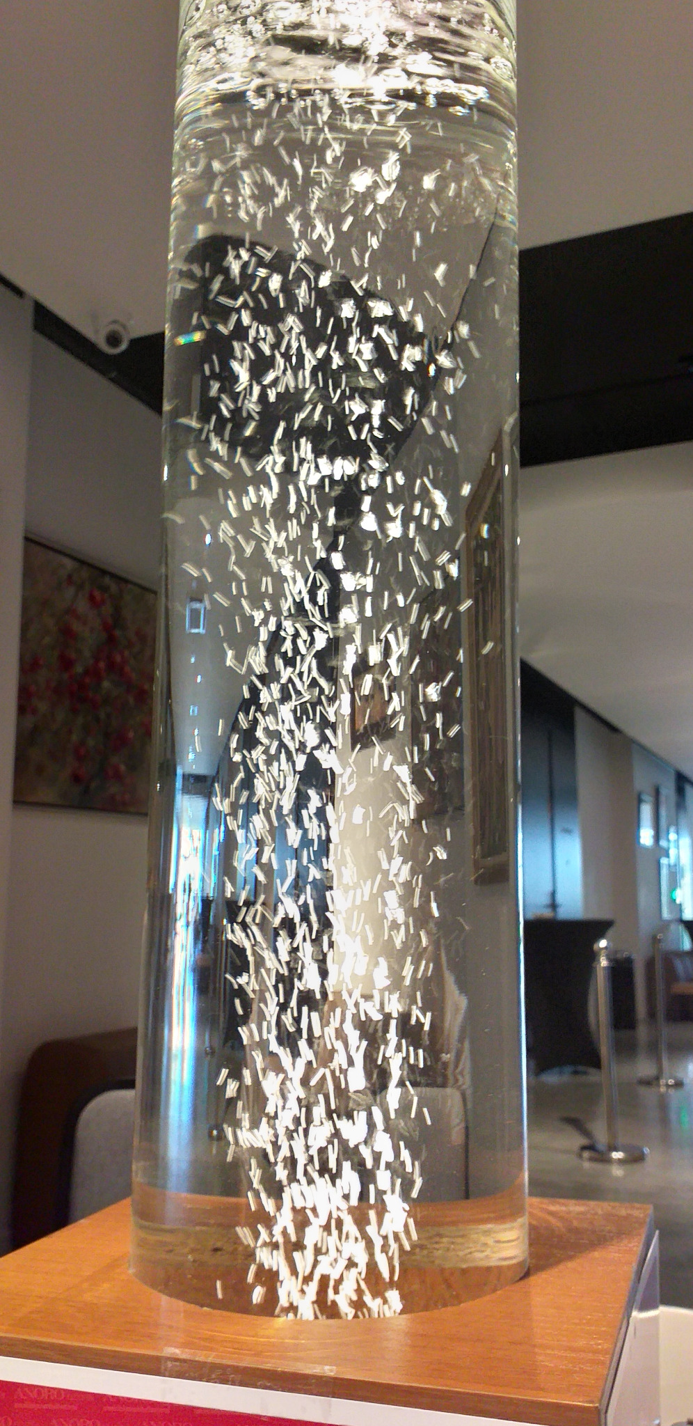 Szklana kolumna wypełniona wodą z unoszącymi się białymi, prostokątnymi elementami, tworzącymi efekt dekoracyjny w przestrzeni publicznej.