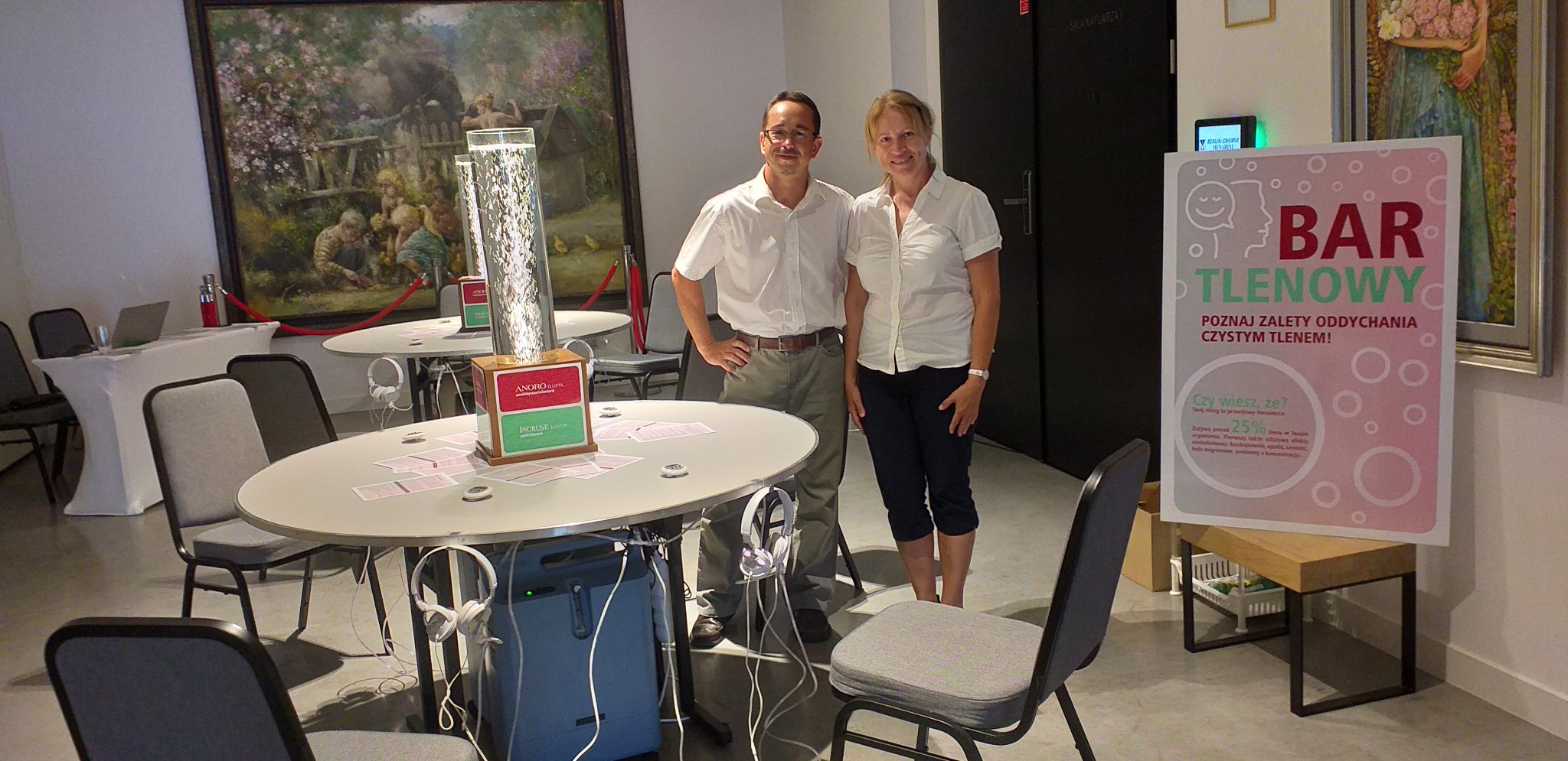 Dwie osoby stoją w pomieszczeniu obok stolików z urządzeniami do inhalacji tlenem, na stolikach leżą słuchawki, w tle obraz i plakat z napisem 'Bar Tlenowy'.