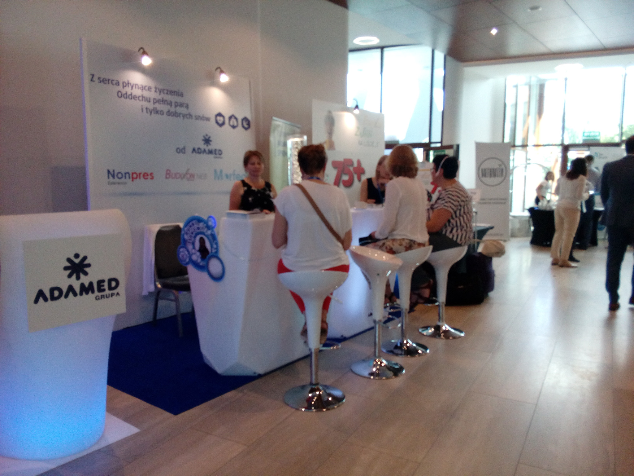 Stoisko firmy Adamed na evencie, białe lady podświetlone na niebiesko, osoby siedzące na wysokich krzesłach, w tle widoczni inni uczestnicy wydarzenia.