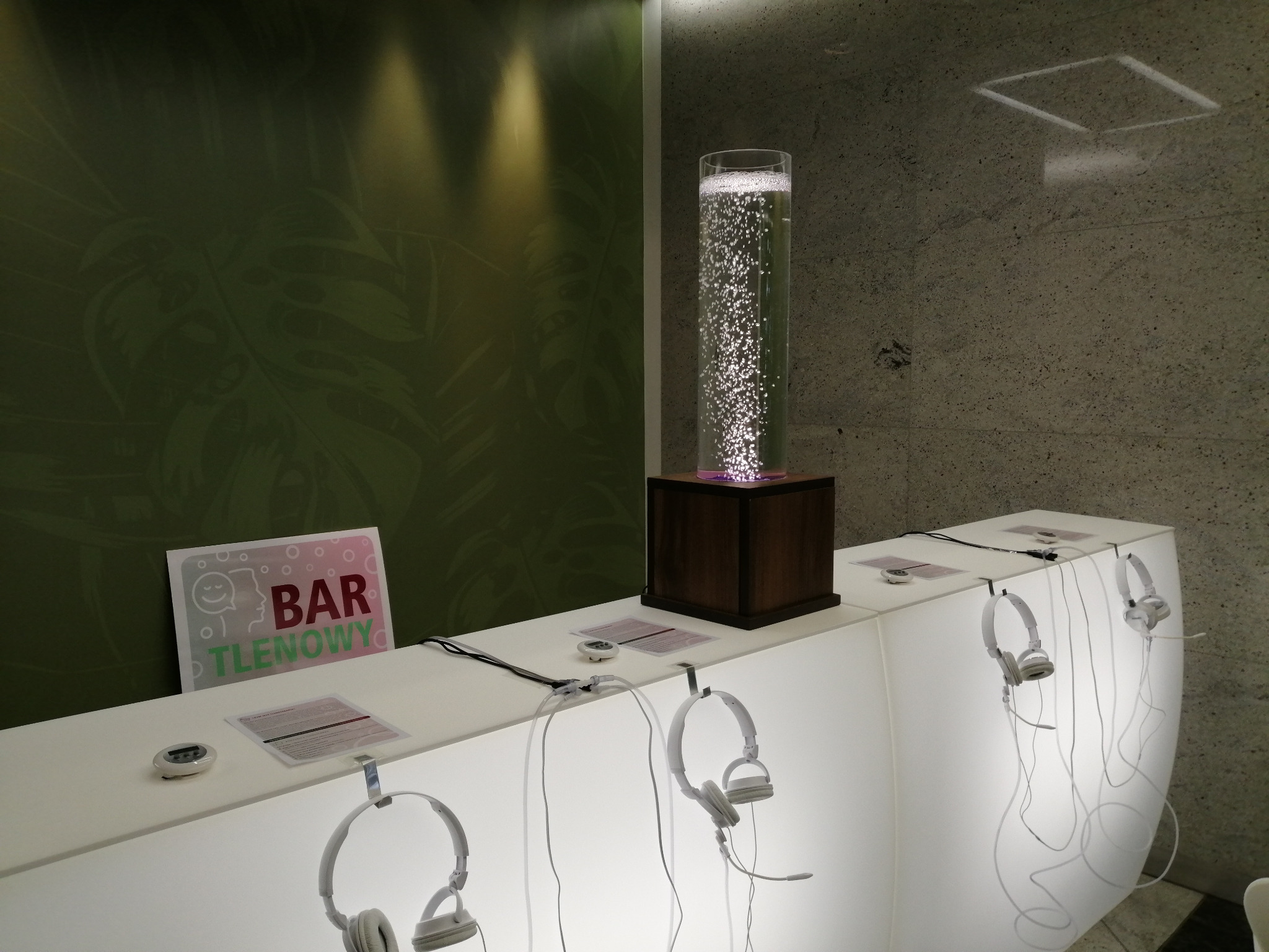 Bar tlenowy z białym, podświetlonym blatem, na którym wiszą słuchawki. W tle zielona ściana z motywem liści i kolumna z bąbelkami powietrza.