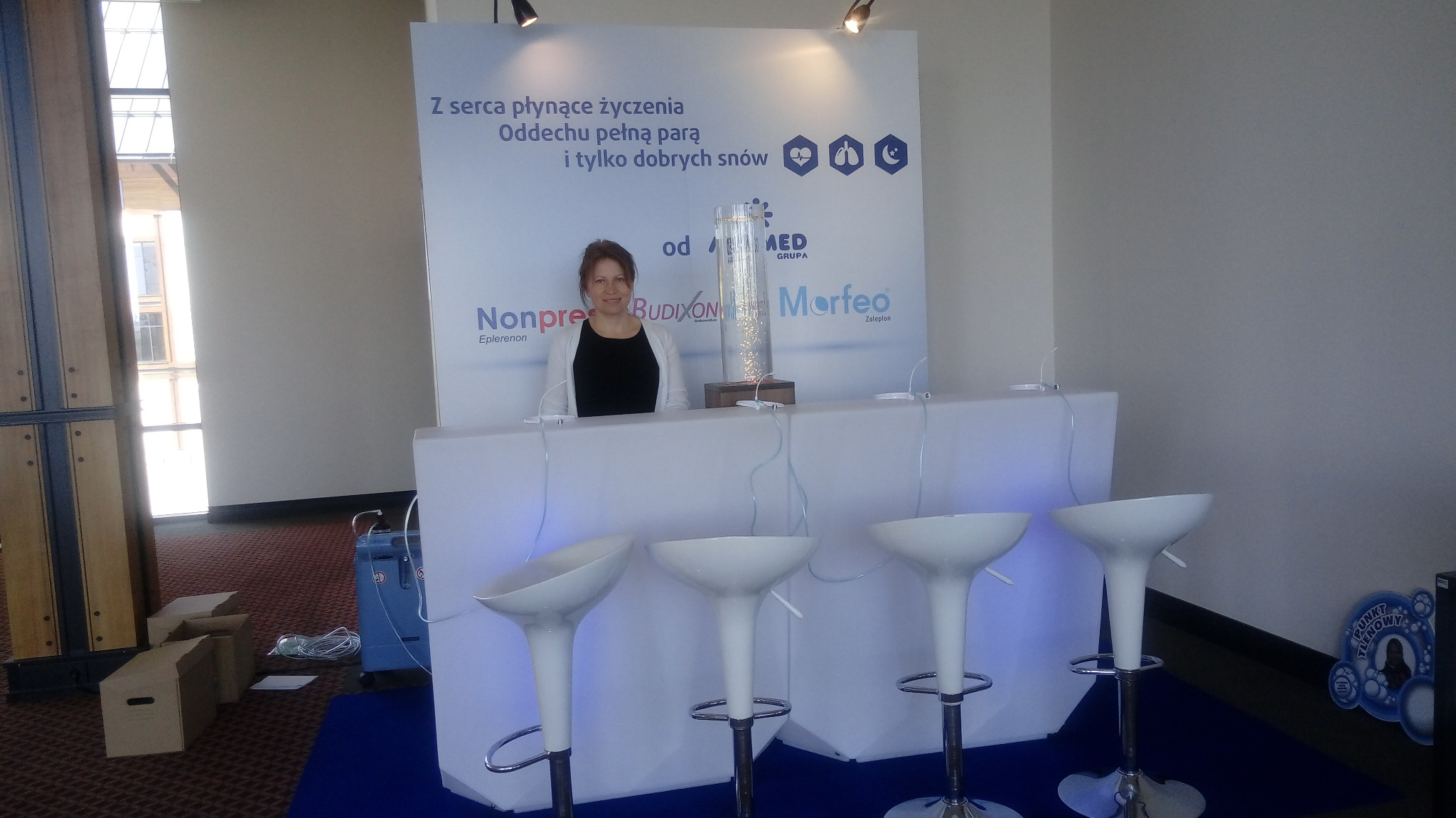 Stoisko promocyjne firmy z Trzebnicy, z podświetlanym blatem, wysokimi białymi krzesłami barowymi i elegancką dekoracją z podświetlaną kolumną wodną oraz uśmiechniętą kobietą za ladą na tle baneru...