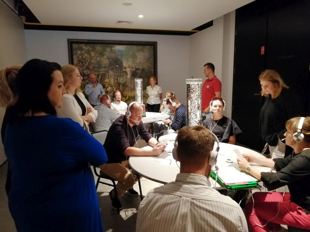 Grupa osób w słuchawkach siedzi przy okrągłych stołach, prawdopodobnie podczas szkolenia lub warsztatów, w tle obraz i kolumna z bąbelkami.