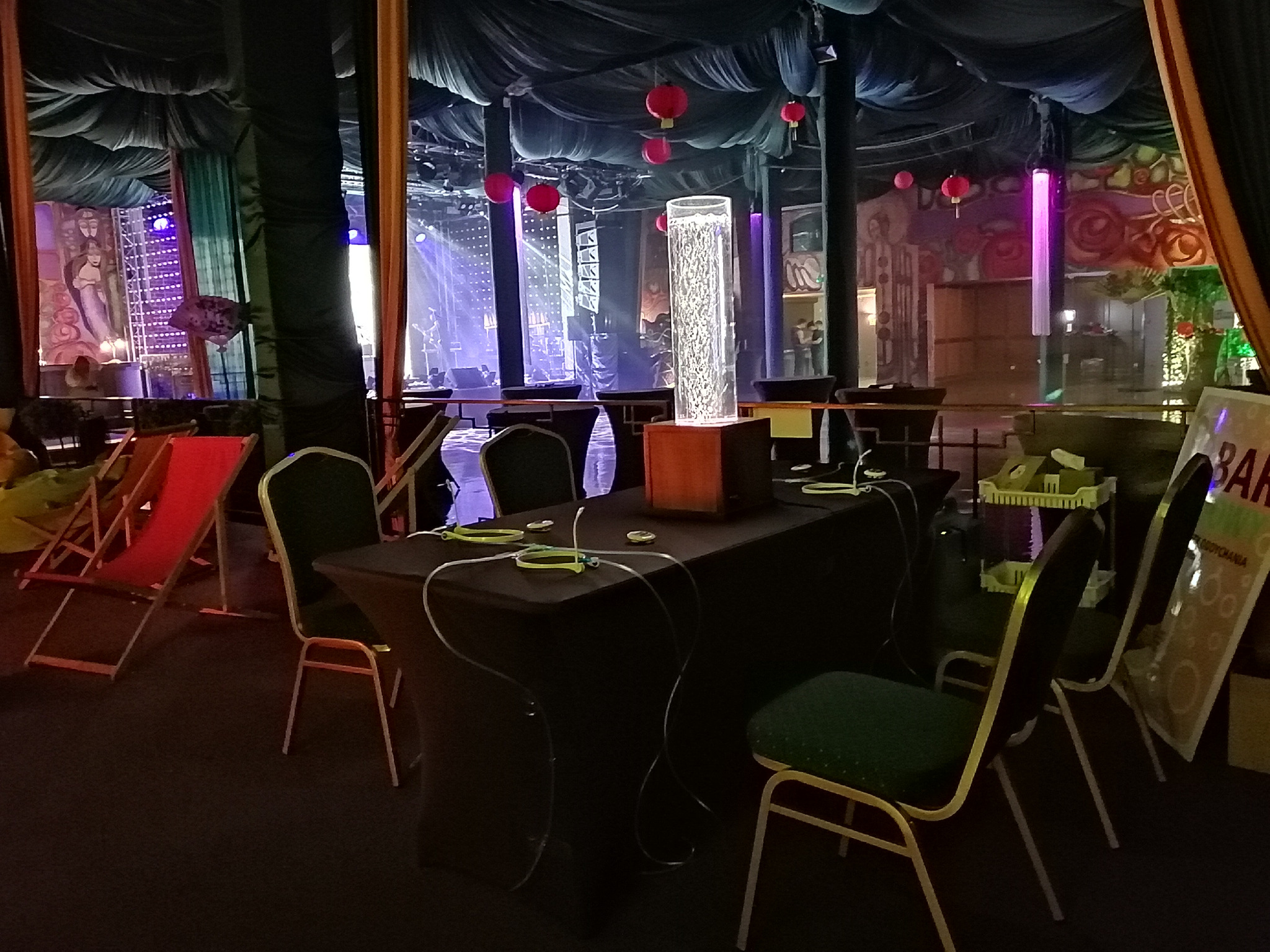 Przygotowania do eventu: stoliki z podświetlanymi dekoracjami, krzesła i leżaki w przestrzeni z dekoracjami, sceną i czerwonymi lampionami.