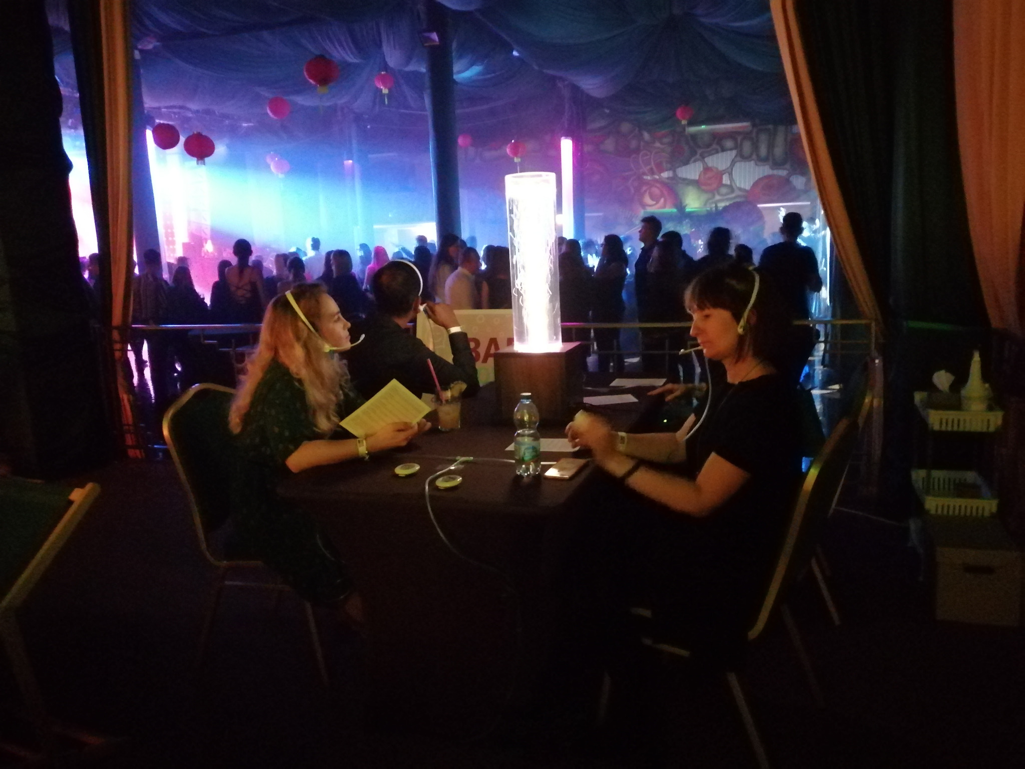 Dwie osoby siedzą przy stole w ciemnym pomieszczeniu z dekoracjami w tle, prawdopodobnie podczas cichej dyskoteki, słuchając muzyki przez słuchawki.