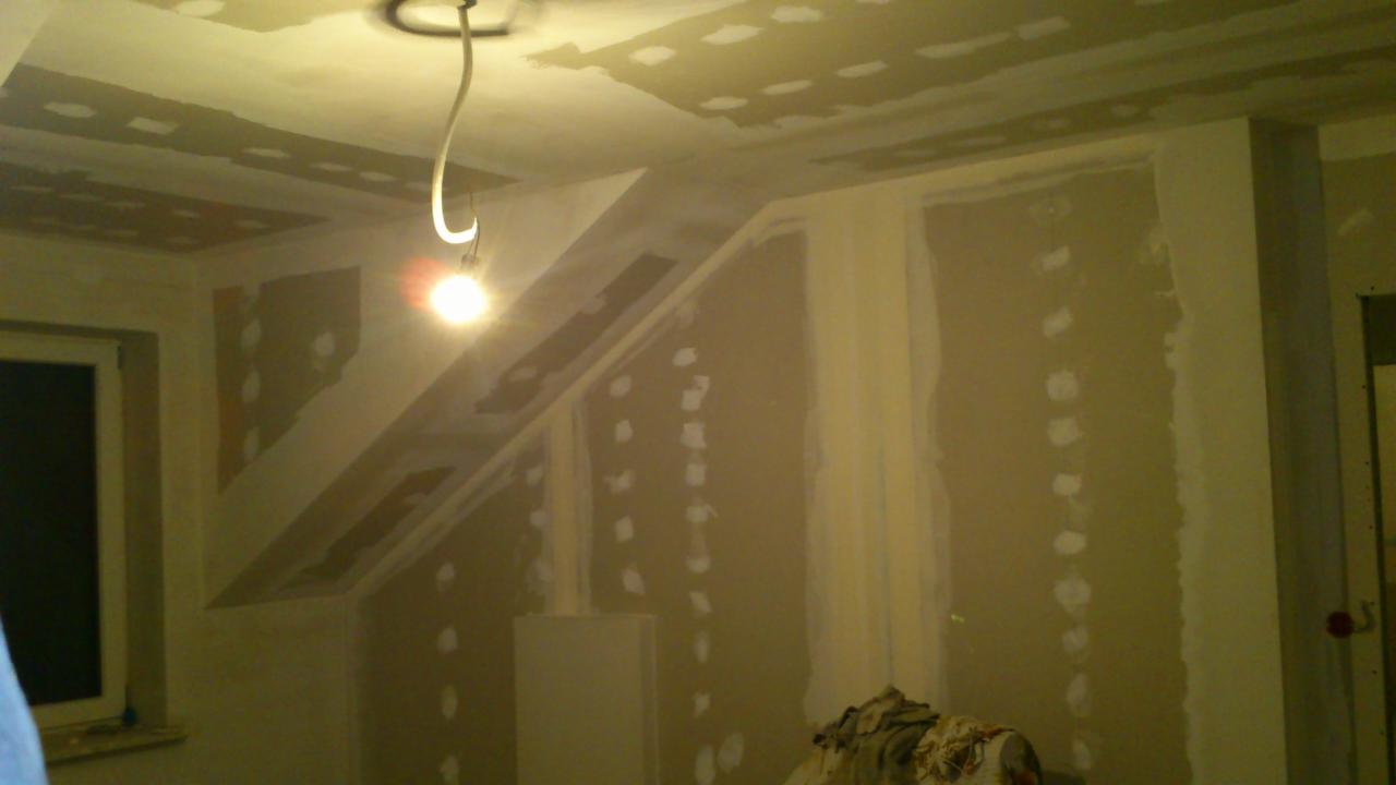 Poddasze w trakcie docieplania, widoczne płyty gipsowo-kartonowe przytwierdzone do skosu dachu, ślady po szpachlowaniu, zwisająca żarówka oświetla wnętrze.