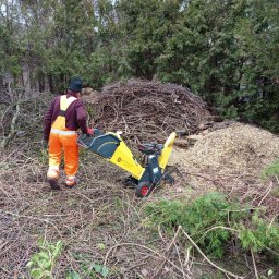 MNM Invest Usługi Ogrodniczo Budowlane Grzegorz Zięba - Pracownik w pomarańczowym kombinezonie prowadzi żółto-czarną rozdrabniarkę gałęzi na tle sterty gałęzi i zrębków.