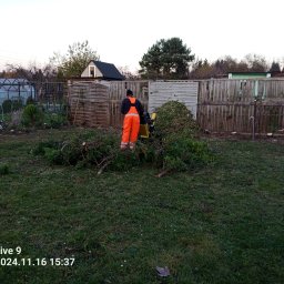 MNM Invest Usługi Ogrodniczo Budowlane Grzegorz Zięba - Pracownik w pomarańczowym kombinezonie obsługuje rozdrabniacz gałęzi na trawniku, w tle drewniany płot i zabudowania gospodarcze.