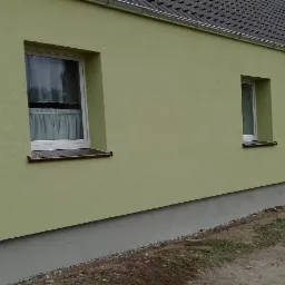 Świeżo pomalowana elewacja domu w odcieniach zieleni, widoczne trzy okna z brązowymi parapetami, szara podmurówka i fragment dachu z rynnami. W tle zabudowa jednorodzinna i pochmurne niebo.