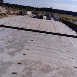 Wylana płyta fundamentowa z drewnianymi elementami dystansowymi, widoczne szalunki i zbrojenia, w tle las i błękitne niebo.