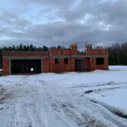 Surowy szkielet domu z czerwonej cegły w zimowej scenerii, widoczne ślady opon na śniegu, las w tle, pochmurne niebo.