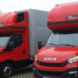 Dwa czerwone samochody dostawcze Iveco z nadbudową typu furgon, zaparkowane na kostce brukowej, z widocznymi numerami rejestracyjnymi i logo 'Sporar' na nadbudowie.