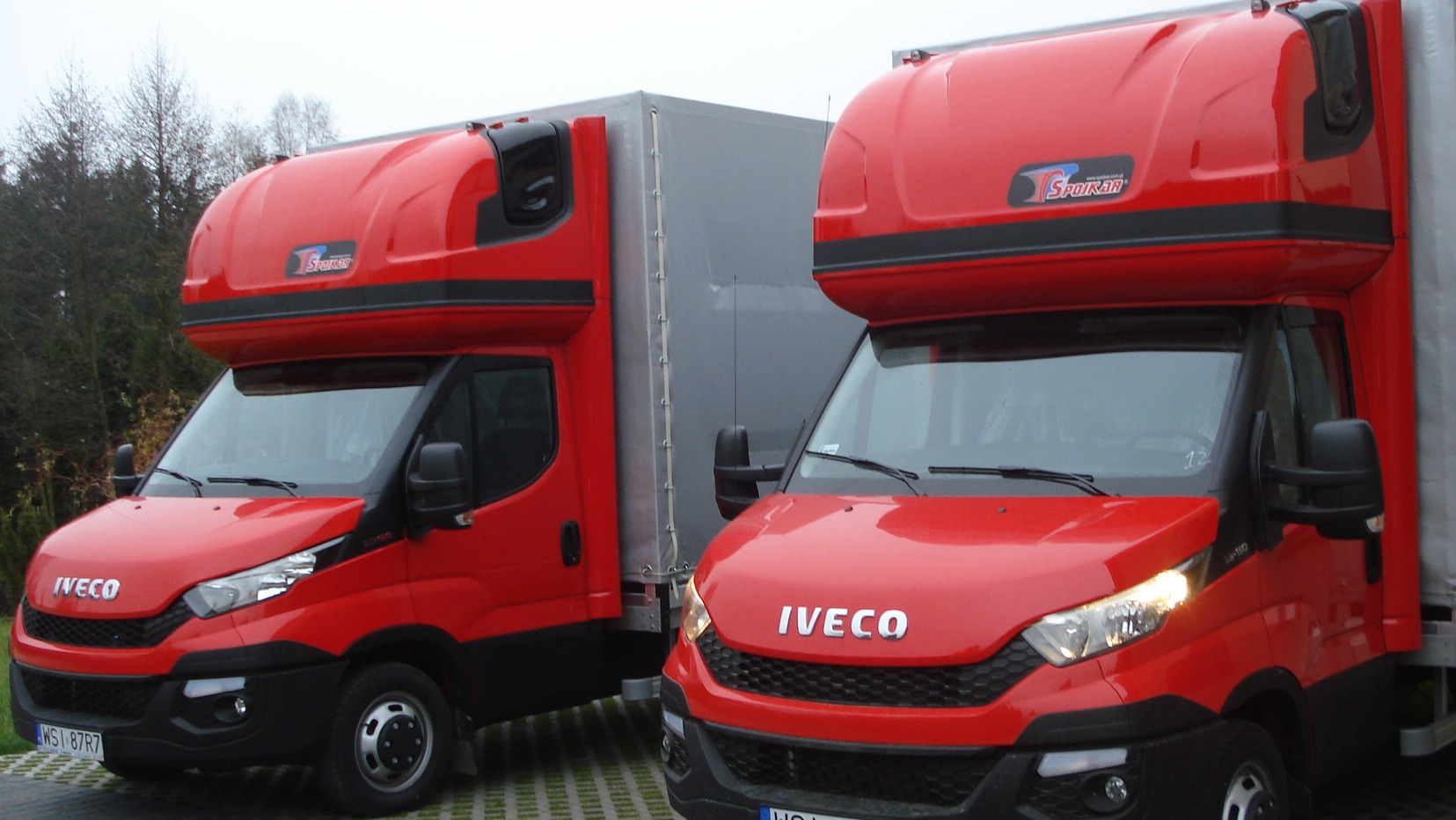 Dwa czerwone samochody dostawcze Iveco z nadbudową typu furgon, zaparkowane na kostce brukowej, z widocznymi numerami rejestracyjnymi i logo 'Sporar' na nadbudowie.