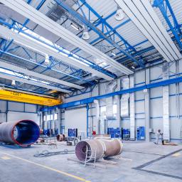 Clima Line Sompoliński Spółka Jawna - Wnętrze hali produkcyjnej z elementami instalacji przemysłowych: duże rury na wózkach, suwnica pod stropem, widoczne kanały wentylacyjne i klimatyzacyjne pod sufitem, pracownik w kombinezonie w tle.