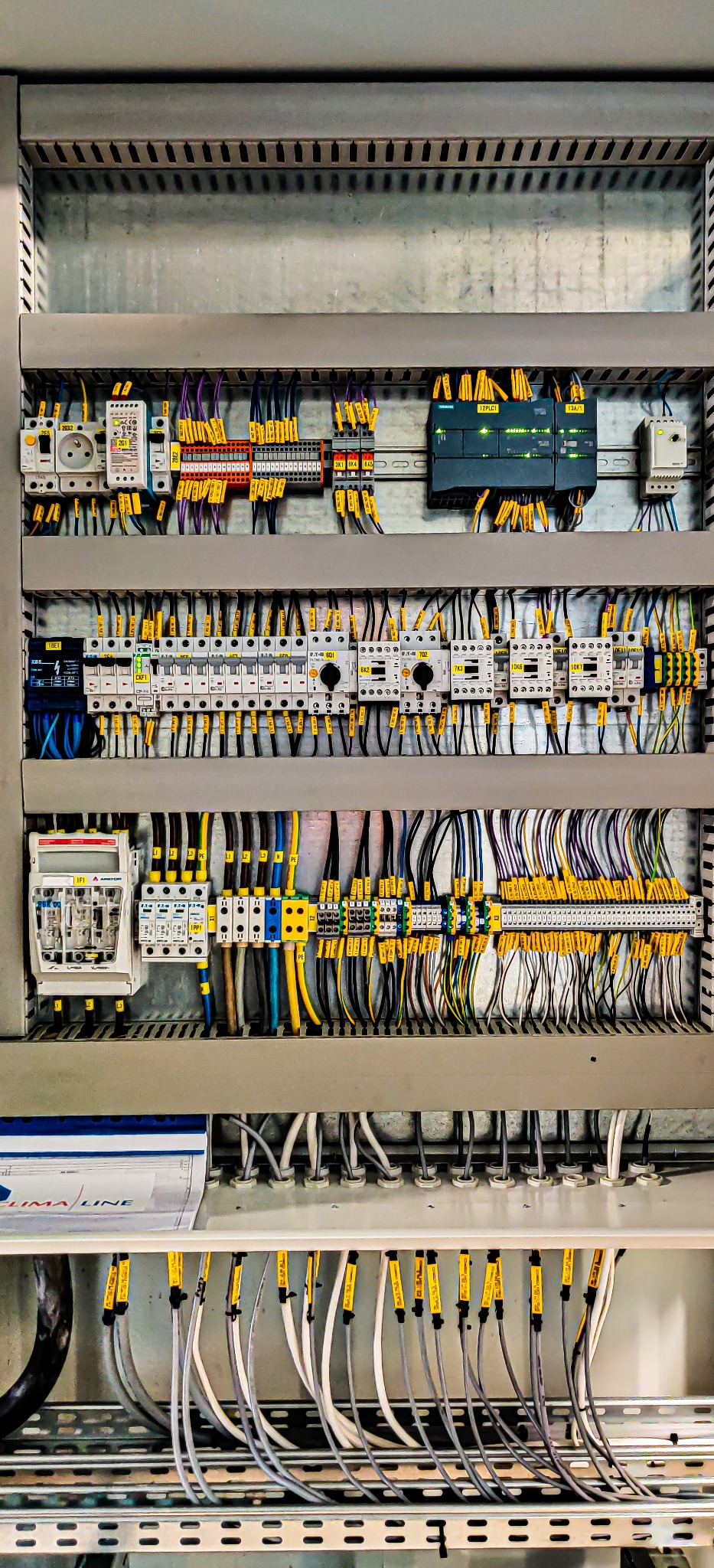 Rozdzielnica elektryczna z licznymi przewodami oznaczonymi żółtymi etykietami, widoczne bezpieczniki, przekaźniki i moduł sterujący Siemens, całość umieszczona w szarej obudowie.