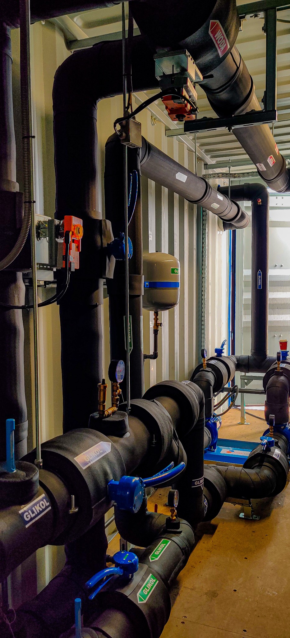 Złożony układ rur w izolacji, z zaworami i manometrami, oznaczony etykietami 'GLIKOL' i 'WODA', zainstalowany w pomieszczeniu o ścianach z blachy falistej.