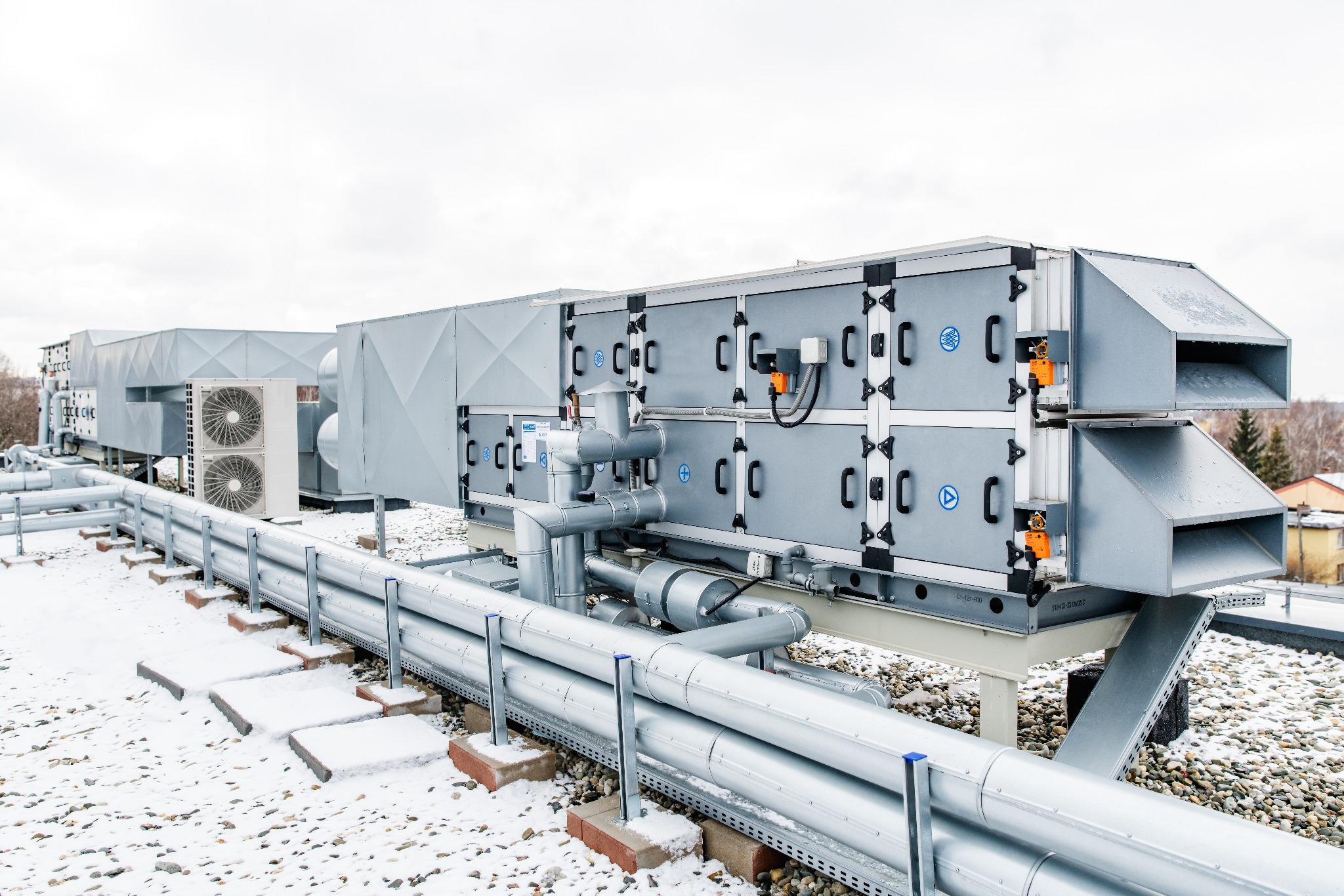 Rozbudowana instalacja wentylacyjna na dachu budynku z widocznymi centralami wentylacyjnymi, rurami i agregatami chłodniczymi, pokryta częściowo śniegiem.