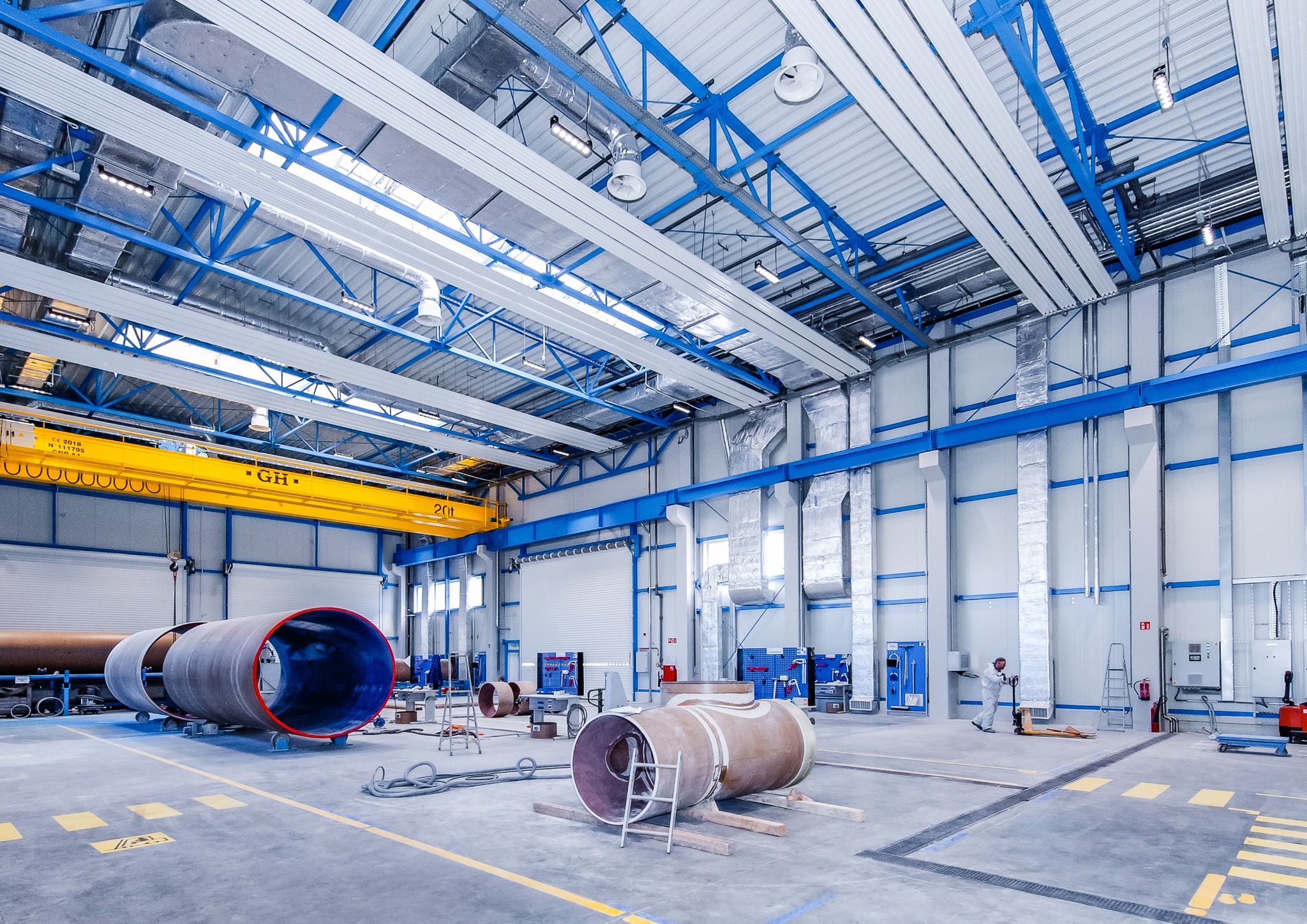 Wnętrze hali produkcyjnej z elementami instalacji przemysłowych: duże rury na wózkach, suwnica pod stropem, widoczne kanały wentylacyjne i klimatyzacyjne pod sufitem, pracownik w kombinezonie w tle.