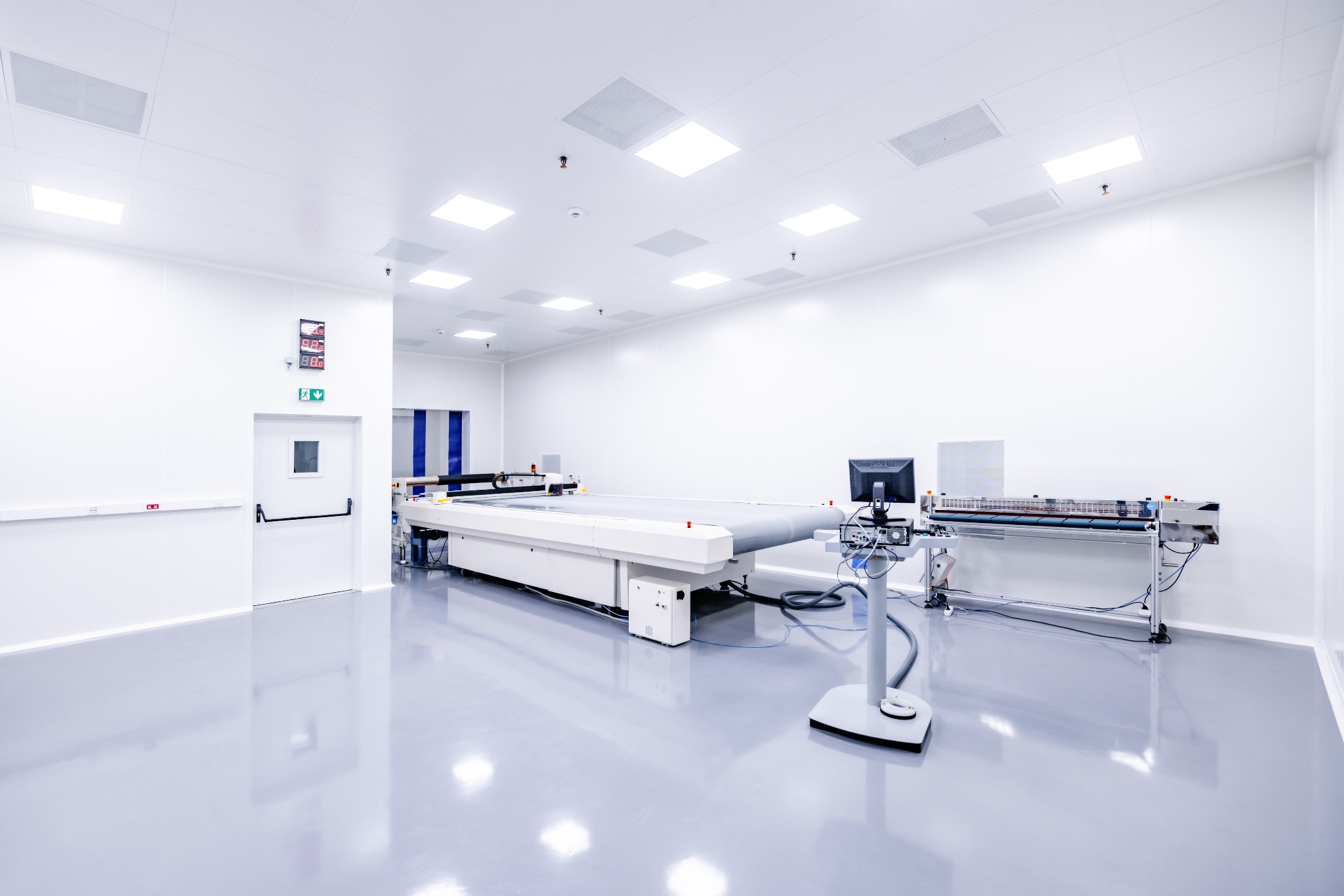 Sterylne pomieszczenie z zaawansowanym sprzętem laboratoryjnym, w tle widoczna tablica informacyjna i drzwi ewakuacyjne.