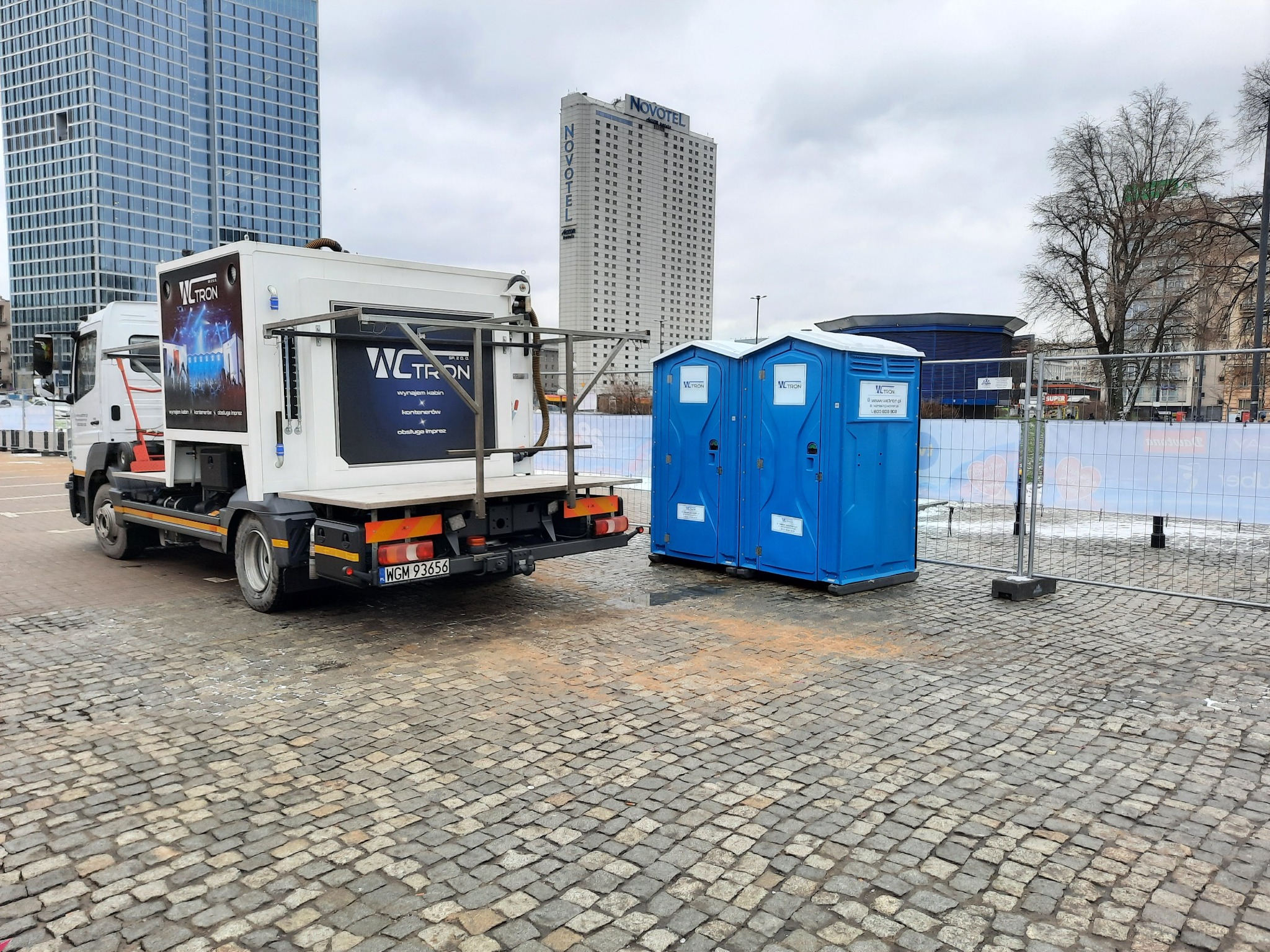 Dwa niebieskie przenośne WC z logo na tle miejskiego krajobrazu z wieżowcami i zaparkowaną ciężarówką z platformą i nadrukowanym logo tej samej firmy.