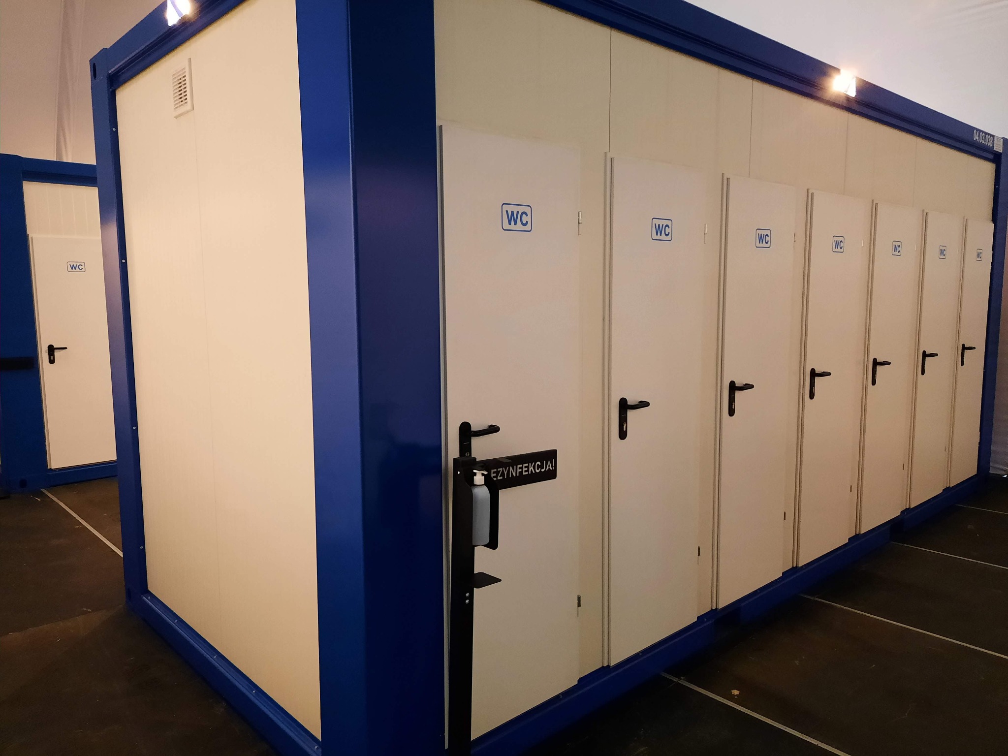 Modułowy kontener sanitarny z rzędem kabin WC, białe drzwi z czarnymi klamkami i oznaczeniami WC, niebieska rama, dozownik do dezynfekcji przy wejściu.