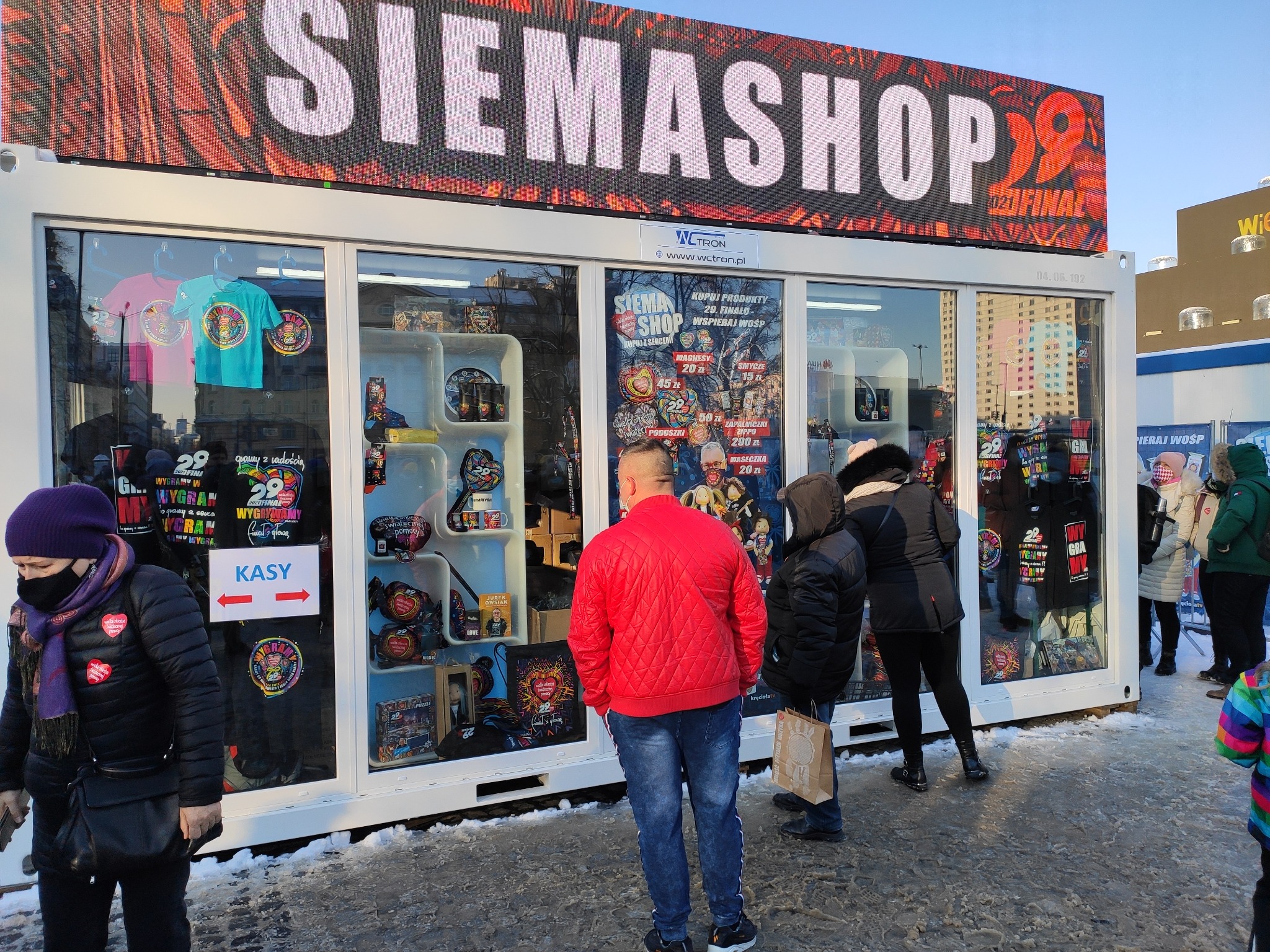 Mobilny sklepik SIEMASHOP z pamiątkami WOŚP, widoczny duży ledowy napis SIEMASHOP 29 Finał, klienci przed witryną, na chodniku resztki śniegu.