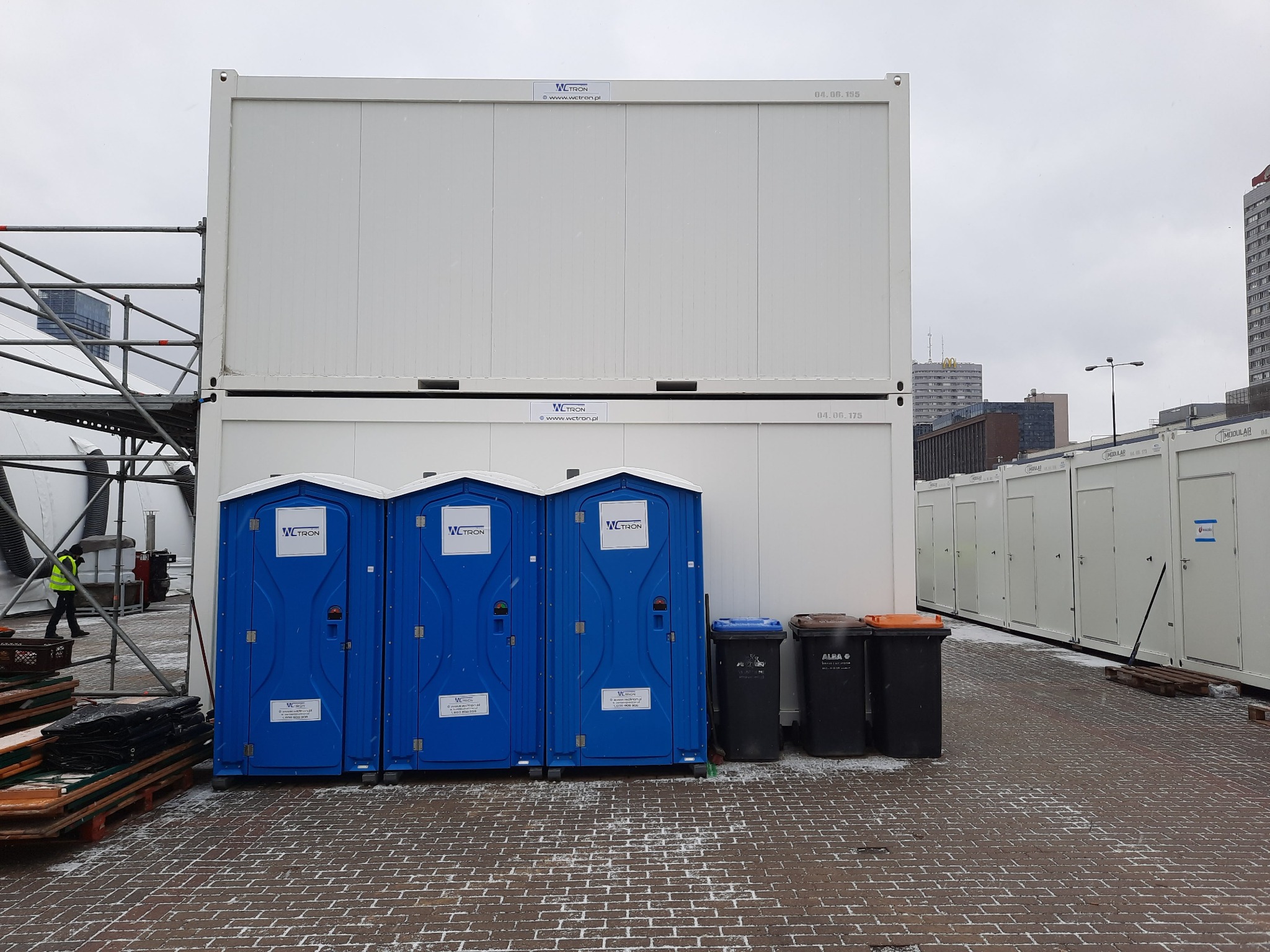 Tymczasowe toalety przenośne i modułowe kontenery biurowe ustawione na placu budowy w pochmurny dzień, widoczne rusztowania i fragment sylwetki miasta w tle.