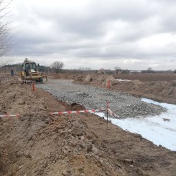 TIRRO Team Robert Traczyk - Żółta koparko-ładowarka CAT rozkłada kamienny tłuczeń na białej geowłókninie, przygotowując podłoże pod drogę na tle pochmurnego nieba i zabudowań.