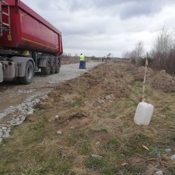 TIRRO Team Robert Traczyk - Ciężarówka wywrotka z ładunkiem kruszywa na budowie drogi w Tarczynie, w tle pracownik zagęszcza podłoże ubijarką, a w oddali widoczny jest sprzęt budowlany.