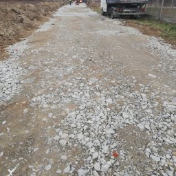 TIRRO Team Robert Traczyk - Wyrównana droga gruntowa z gruzem, w tle pracownik obsługujący zagęszczarkę i wywrotka, po prawej stronie zaparkowany samochód dostawczy.