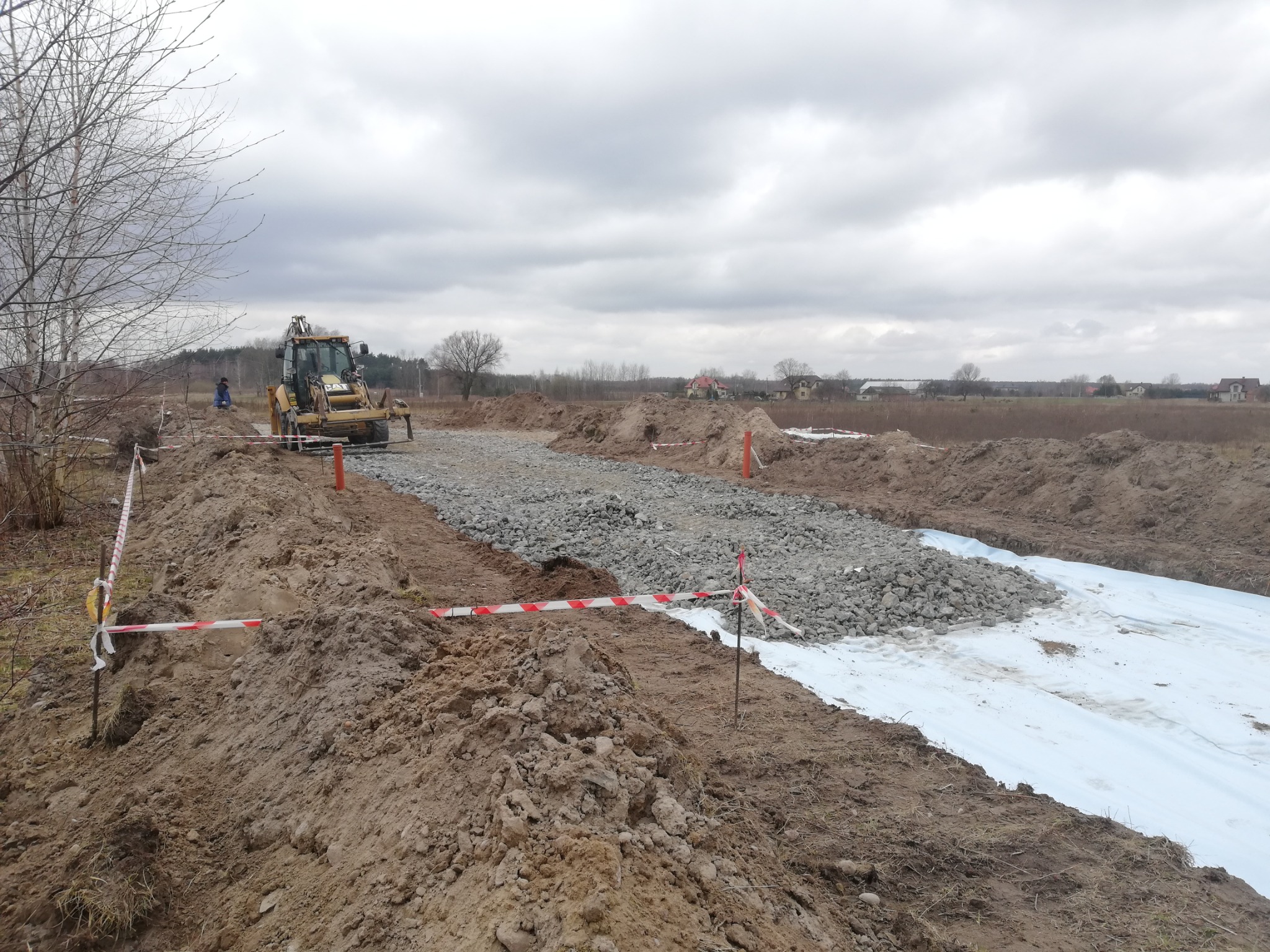 Żółta koparko-ładowarka CAT rozkłada kamienny tłuczeń na białej geowłókninie, przygotowując podłoże pod drogę na tle pochmurnego nieba i zabudowań.