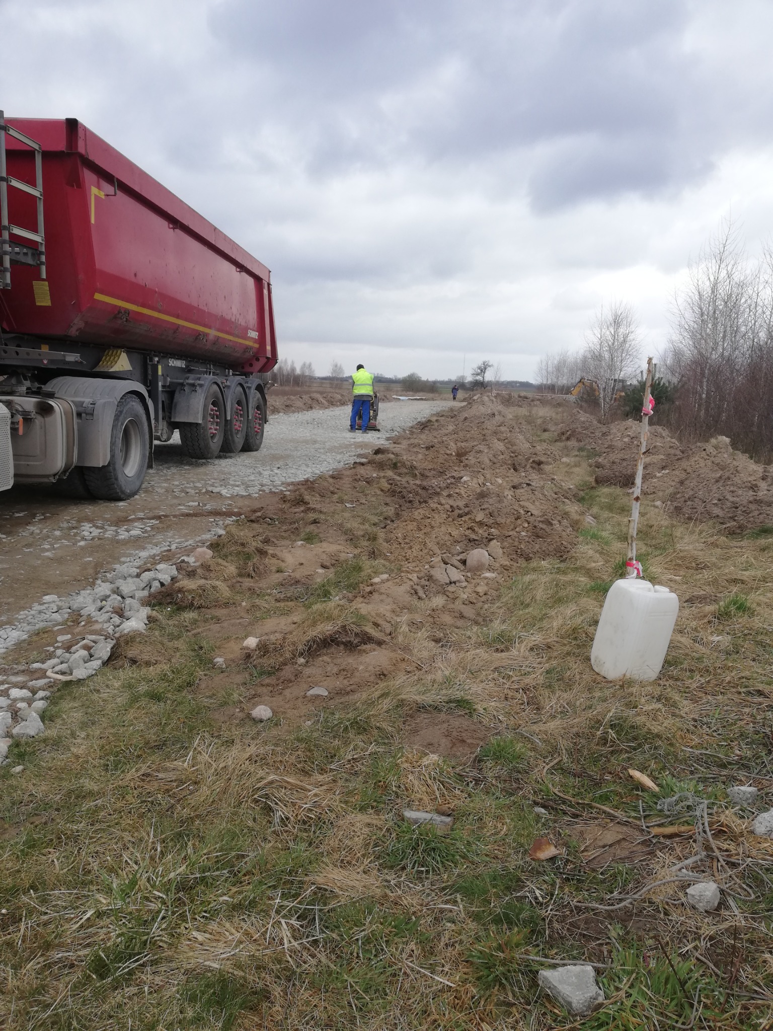 Ciężarówka wywrotka z ładunkiem kruszywa na budowie drogi w Tarczynie, w tle pracownik zagęszcza podłoże ubijarką, a w oddali widoczny jest sprzęt budowlany.