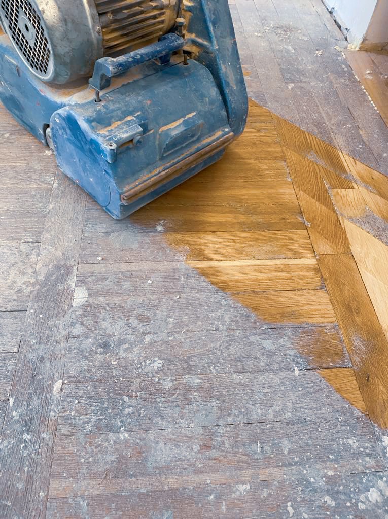 Parkiet w trakcie renowacji z widoczną maszyną do cyklinowania; część podłogi wycyklinowana, ukazująca naturalny kolor drewna, a część pokryta starą warstwą wykończeniową i pyłem.