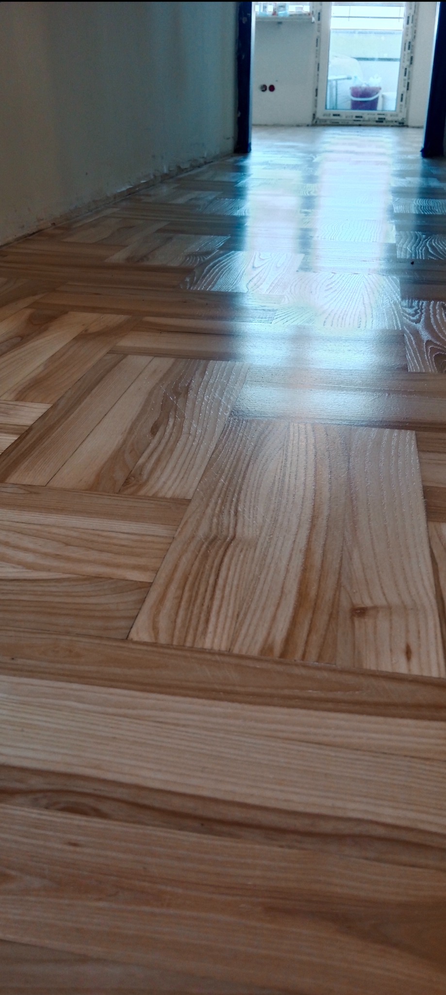 Świeżo położony, lakierowany parkiet w jodełkę, z widocznym odbiciem światła od okna, podkreślającym gładkość powierzchni.