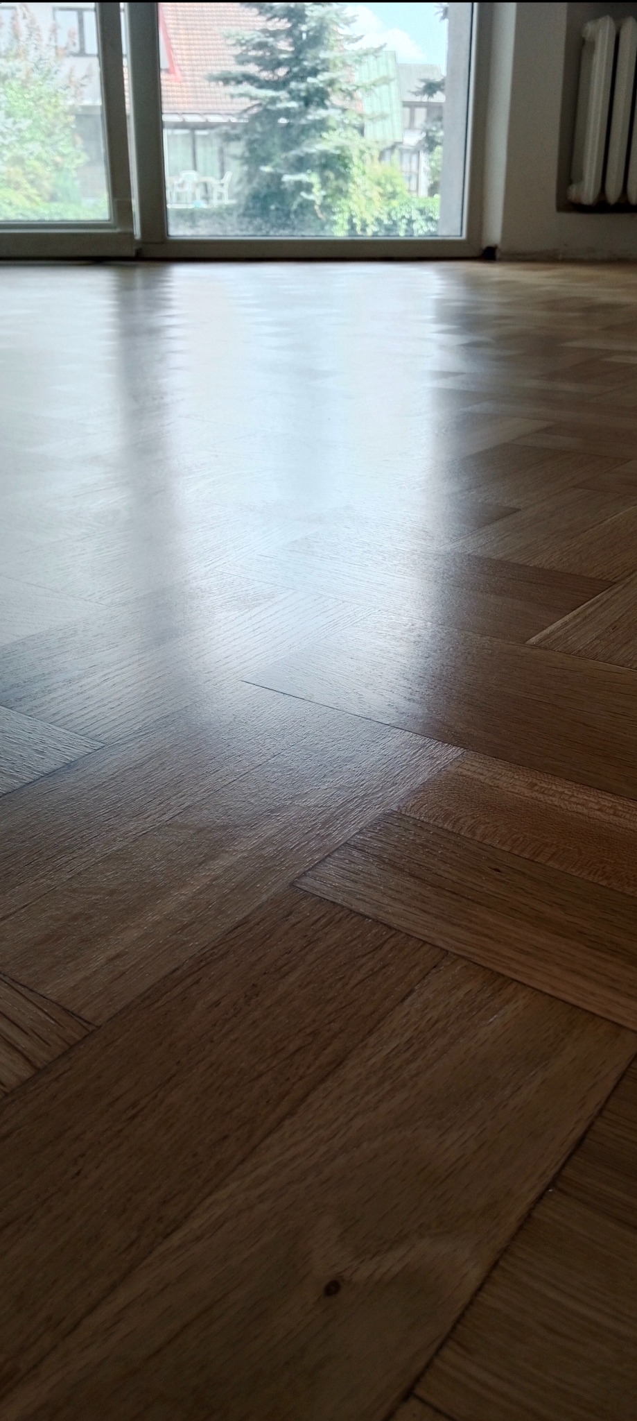 Błyszcząca, świeżo wycyklinowana drewniana podłoga ułożona w jodełkę, odbijająca światło okna z widokiem na zieleń.