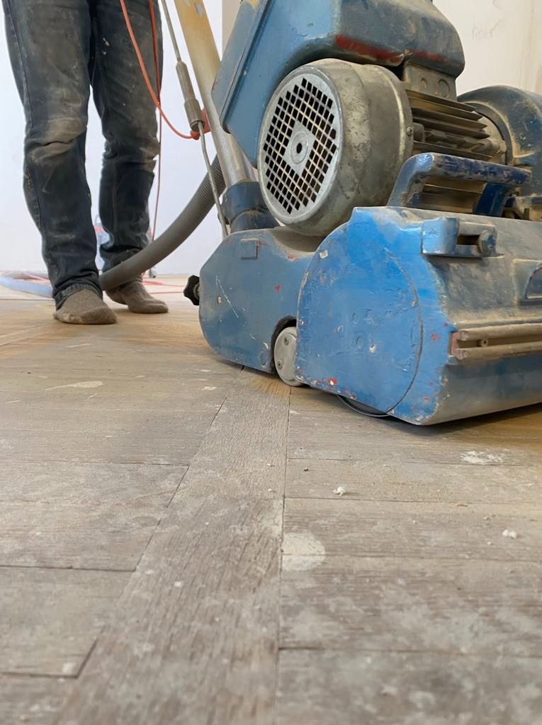 Parkieciarz w trakcie cyklinowania podłogi za pomocą niebieskiej, profesjonalnej szlifierki do parkietu. Widoczne ślady pyłu i niedoskonałości na powierzchni.