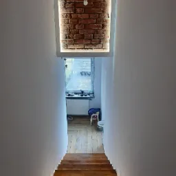 Wnętrze klatki schodowej z drewnianymi schodami, oświetleniem LED pod stopniami i dekoracyjną ścianą z cegły podświetloną lampą wiszącą.